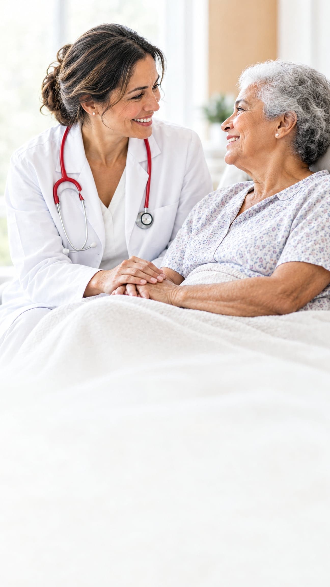 Médica de jaleco branco e estetoscópio vermelho segura as mãos de uma paciente idosa, as duas sorrindo
